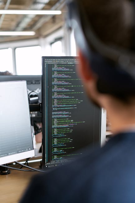 The image shows a person working at a desk in an office environment, viewed from behind. The person has short hair and is wearing glasses and a dark shirt. They are focused on a computer monitor displ