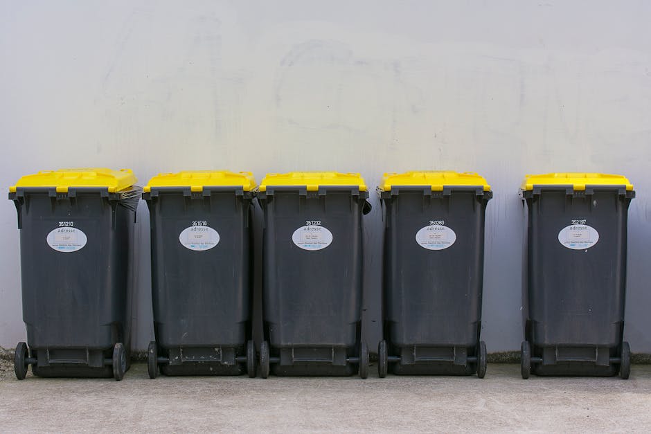 A waste collection operative dressed in an orange safety vest and dark trousers is seen loading rubbish into the rear of a large red refuse collection vehicle parked on a roadside. The back of the veh